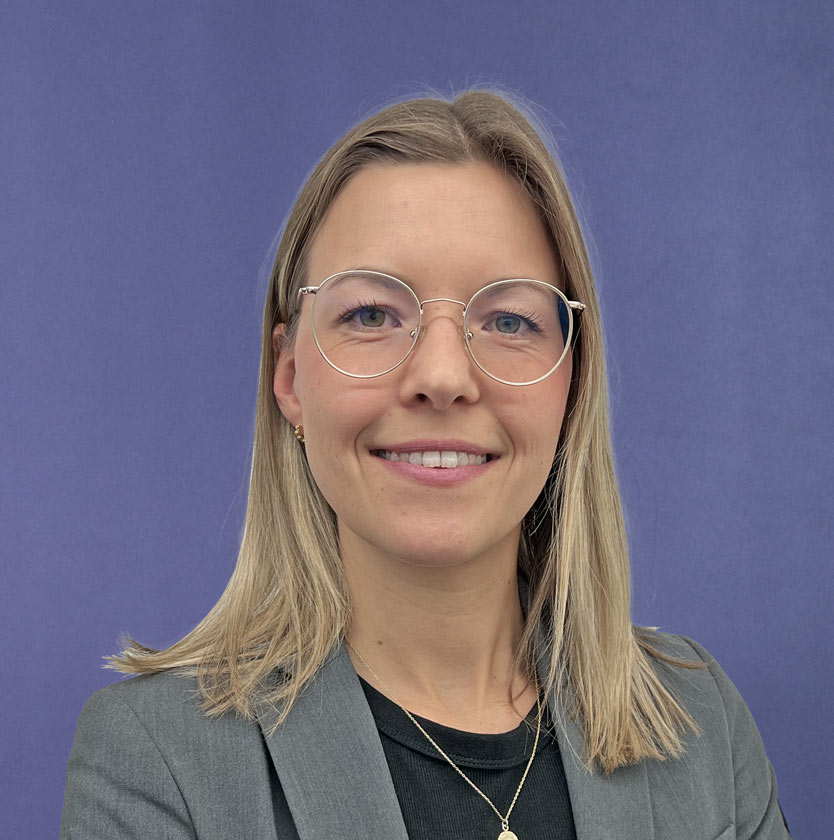 Frau mit runder Brille, grauem Blazer vor blauem Hintergrund.